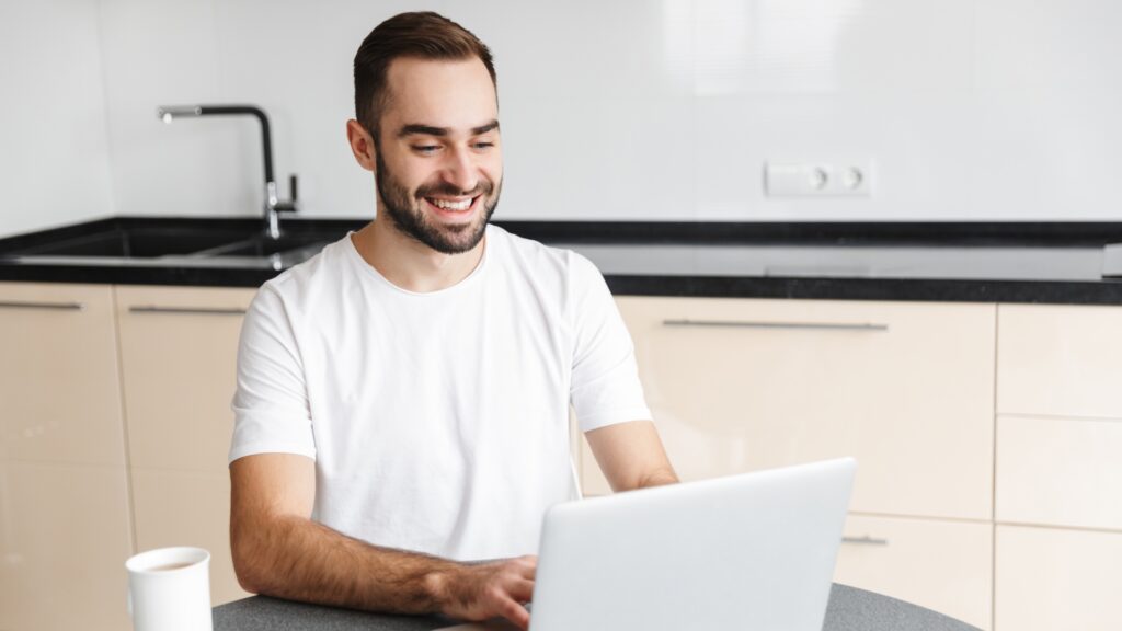 A man in a white T-shirt sits at a desk reading an article on his laptop about the benefits of TransferGo vs Wise Business