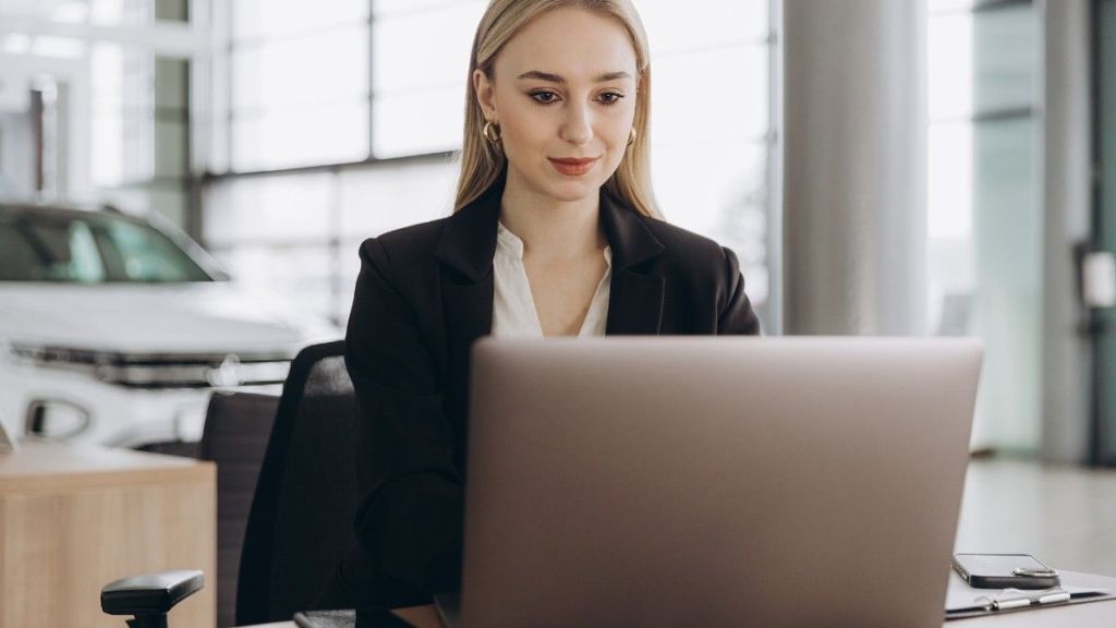 A young blonde female in a black blazer faces the camera as she sits at a desk and types on her laptop about how to manage business expenses