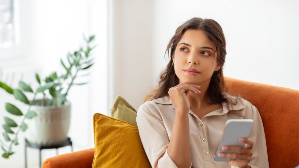 A young female with long brown hair rests her chin on her hand as she sits on an orange sofa and reads an article on her phone that answers the question, 'Is Payoneer safe?'