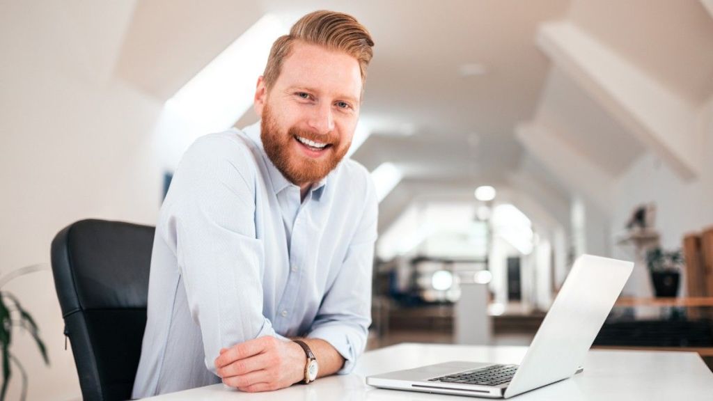 A young man with a beard wears a light shirt and sits at his office desk with his laptop while looking at the camera and smiling as he considers, 'Is Skrill Business safe?'