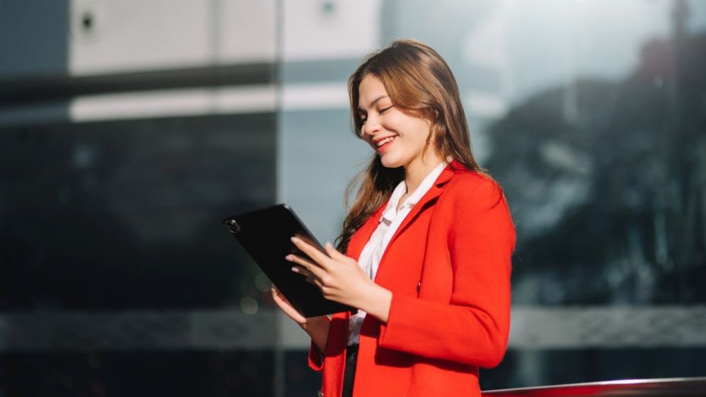 A young brunette female in a red suit stands outside and smiles as she reads an article on her ipad about 'Is Wise Business safe?'