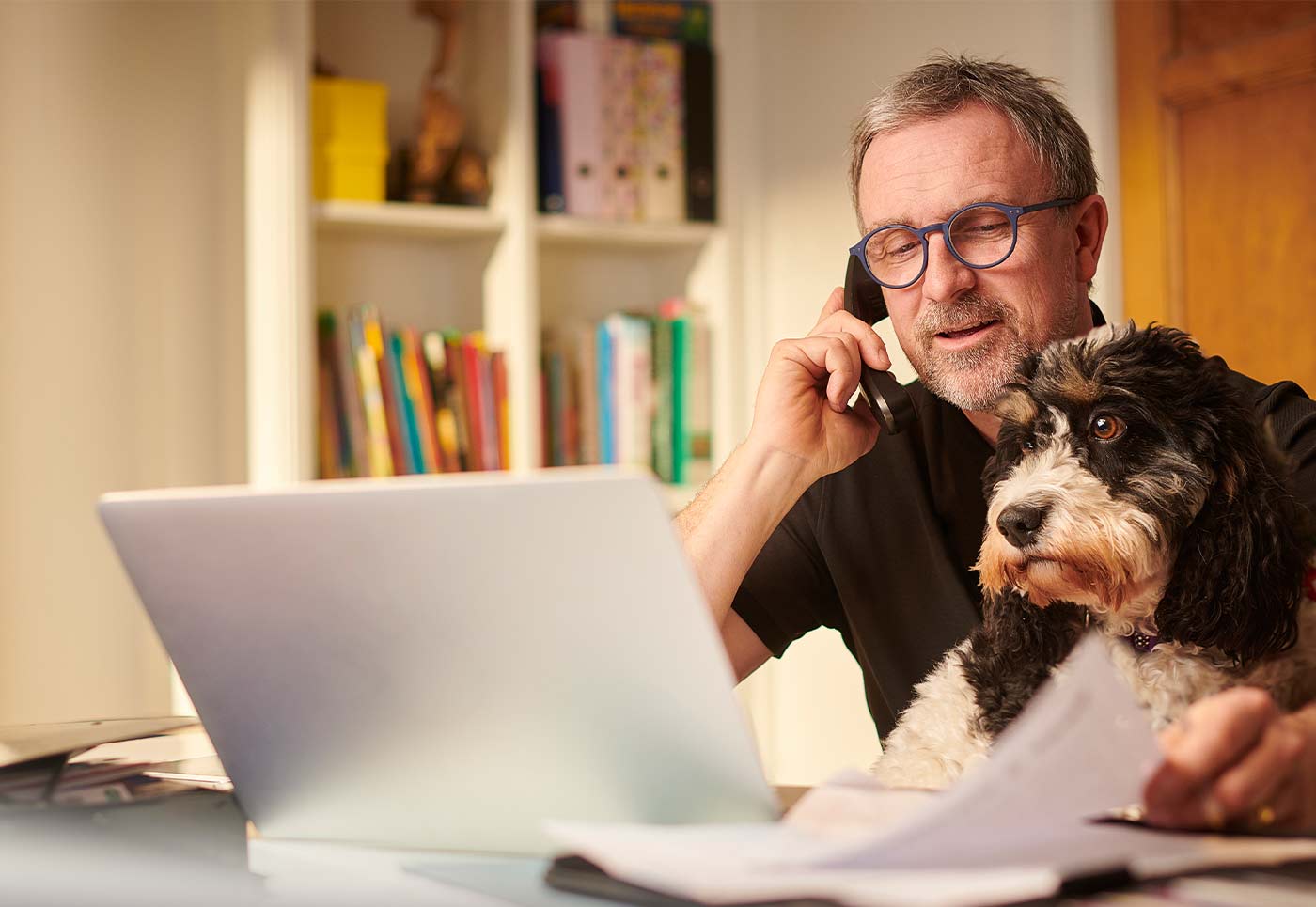 Un bărbat care lucrează la laptop, vorbind la telefon, alături de câinele său, într-un birou de acasă.