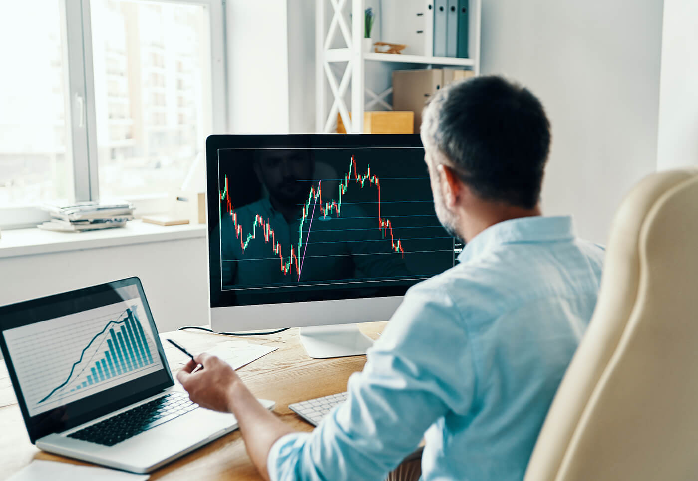 A man in a blue shirt sits at a desk with his back to the camera, looking at a laptop and desktop computer after reading an article about how to become sole trader in the UK