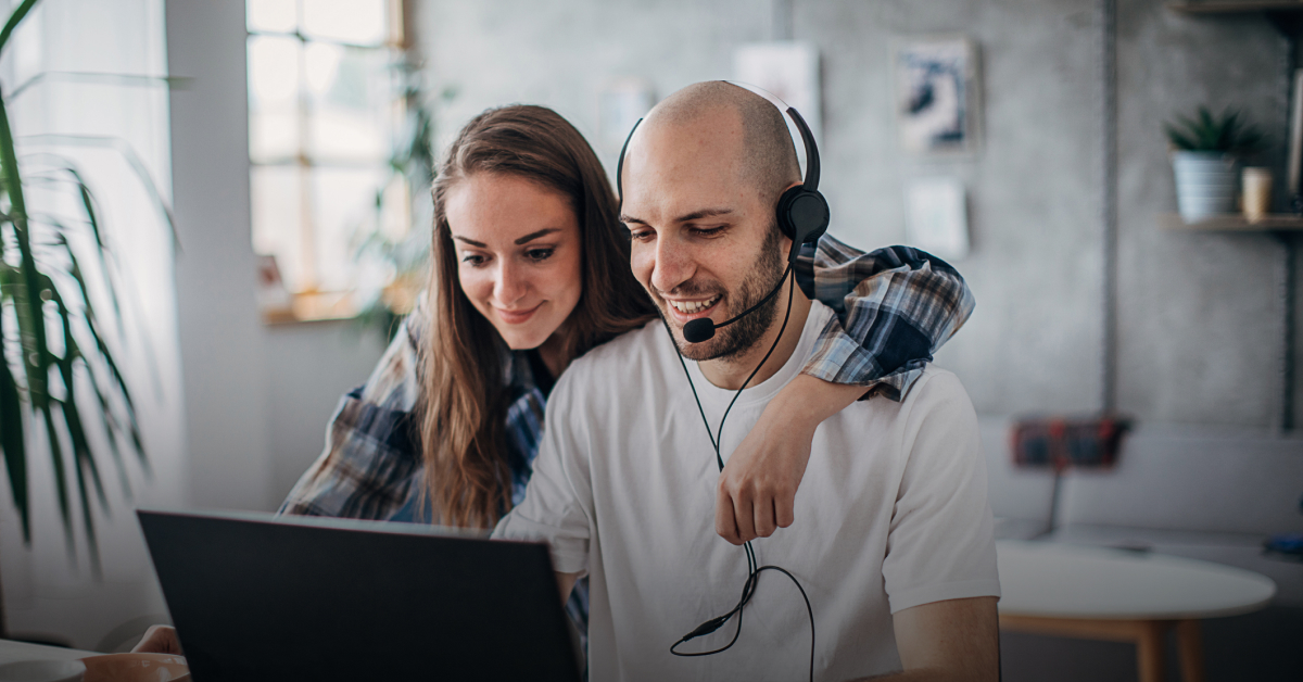 A woman and a man wearing headphones look at a computer. The woman has her arm around the man's shoulders