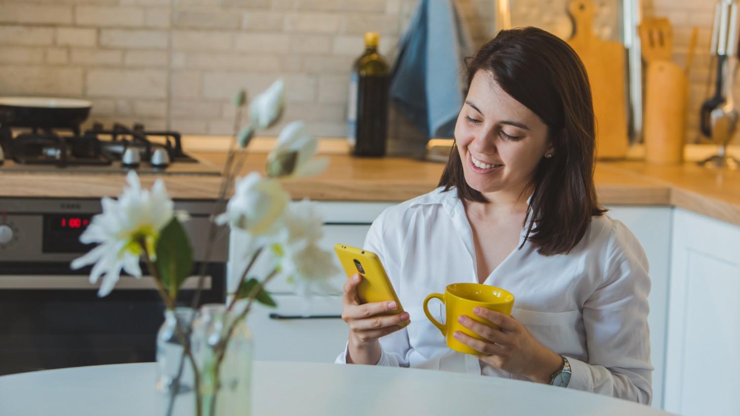 A smiling brunette female sits at the table in her kitchen, reading an article on her phone about the Wise business account