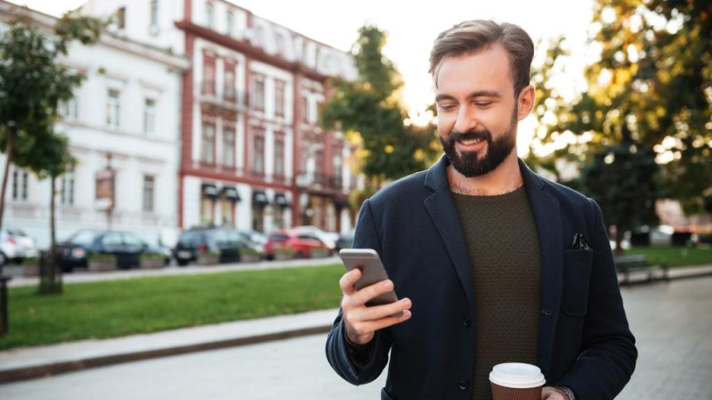 A young bearded male in a grey T-shirt and blue blazer stands outside next to some trees and buildings and looks at his phone, reading an article about the TransferGo BLIK article