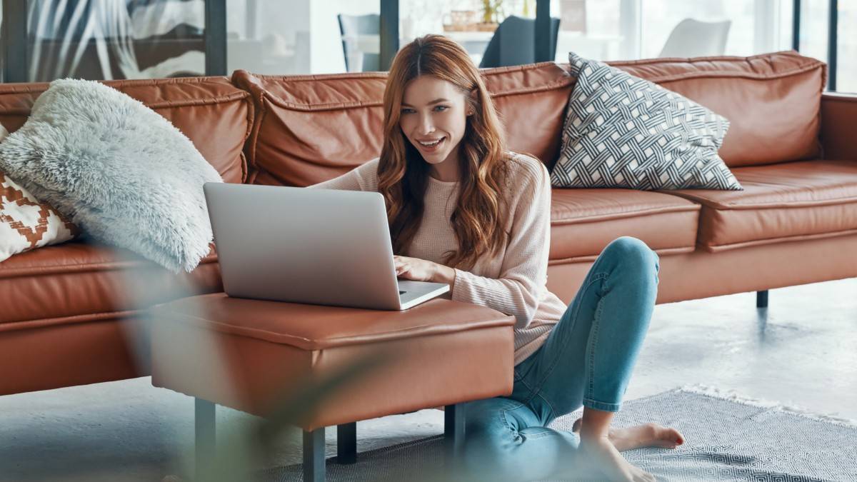 A young female with long red hair sits on the floor in her living room, and rests her laptop on the sofa's foot stool as she reads an article about the TransferGo BLIK partnership