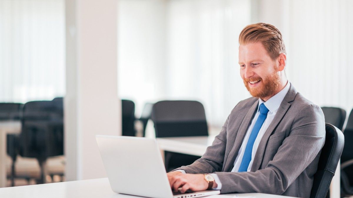 A red-haired man with a beard wears a grey suit and sits at a desk while typing on his laptop and managing business expenses