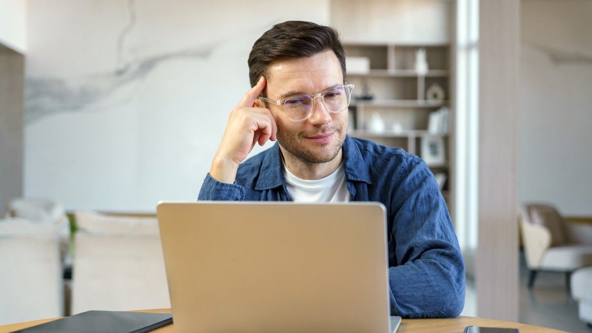 A young man wears a blue shirt and glasses and sits at his desk looking at his laptop and wondering, 'Is Payoneer safe?'