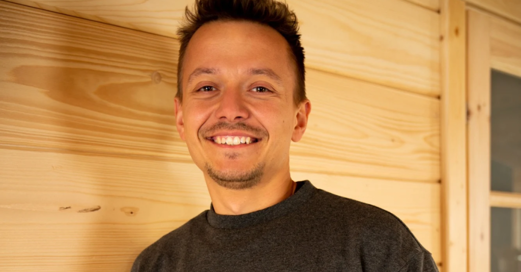 A young man, who is the founder of BaleiDetale.pl, smiles as he wears a dark jumper and stands in front of a wooden wall and door structure