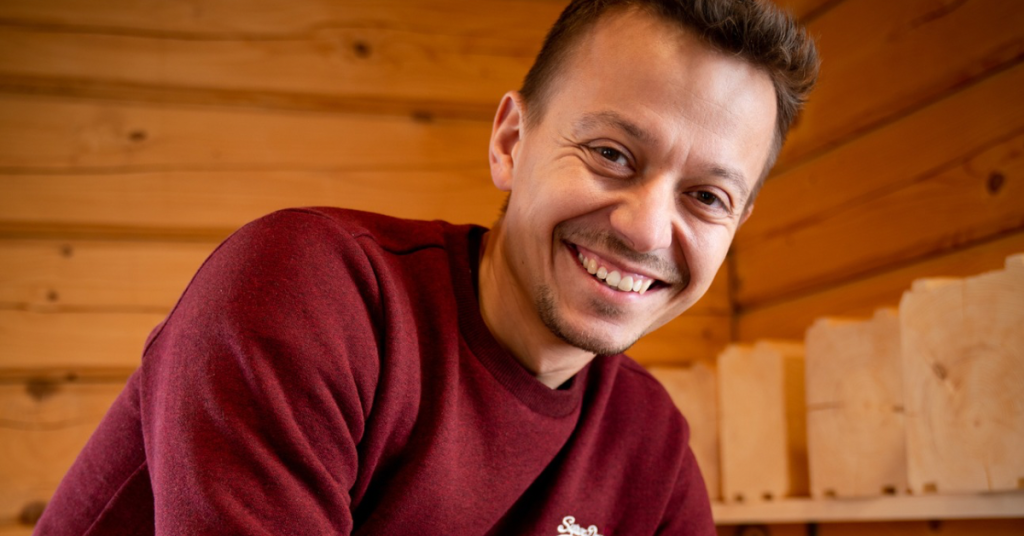 A young man, who is the founder of BaleiDetale.pl, smiles as he wears a red jumper and stands in front of a wooden fence surrounding