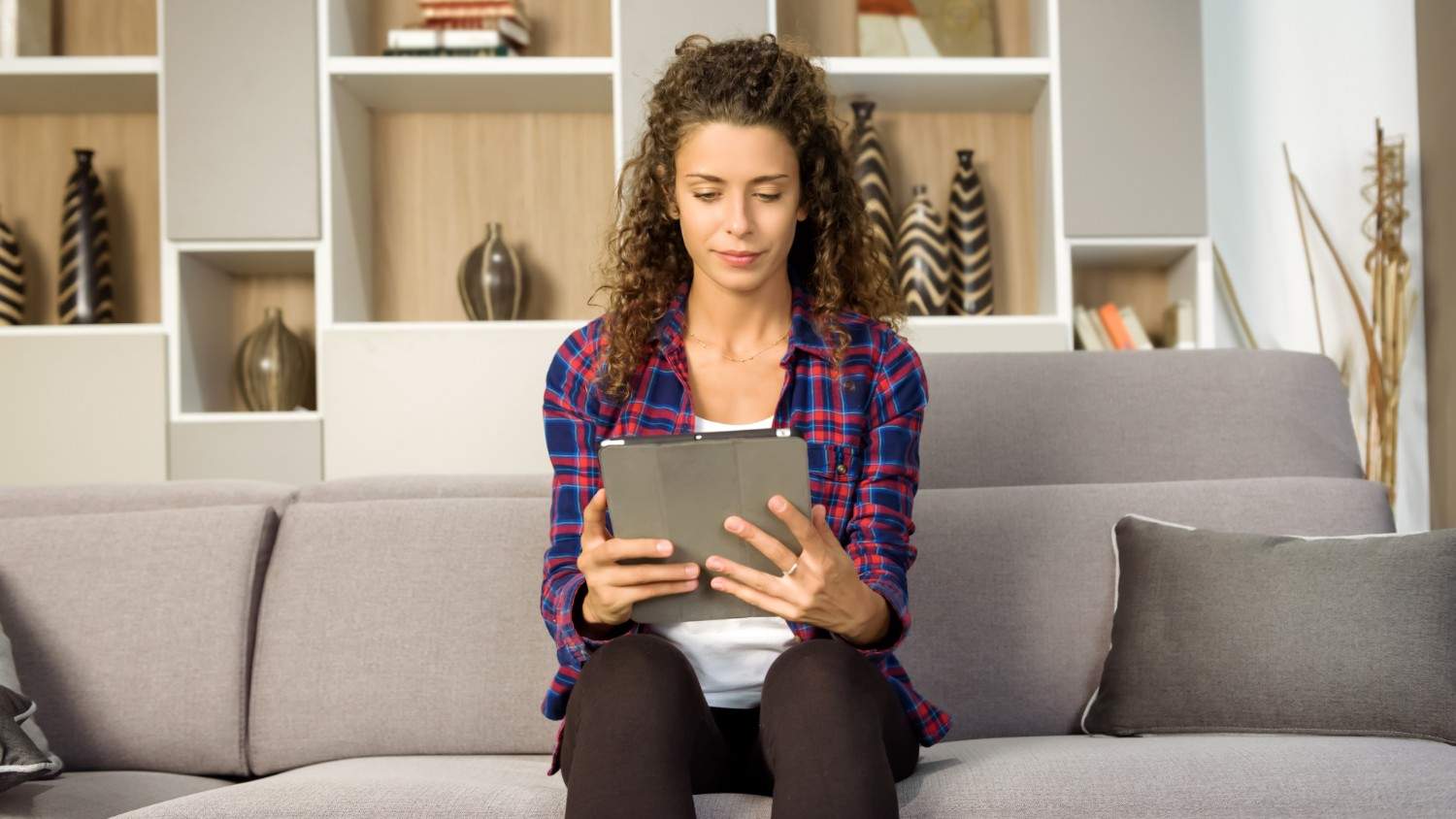 A young brunette female in a checked shirt sits on a sofa and reads an article on her device about how to pay international employees quickly