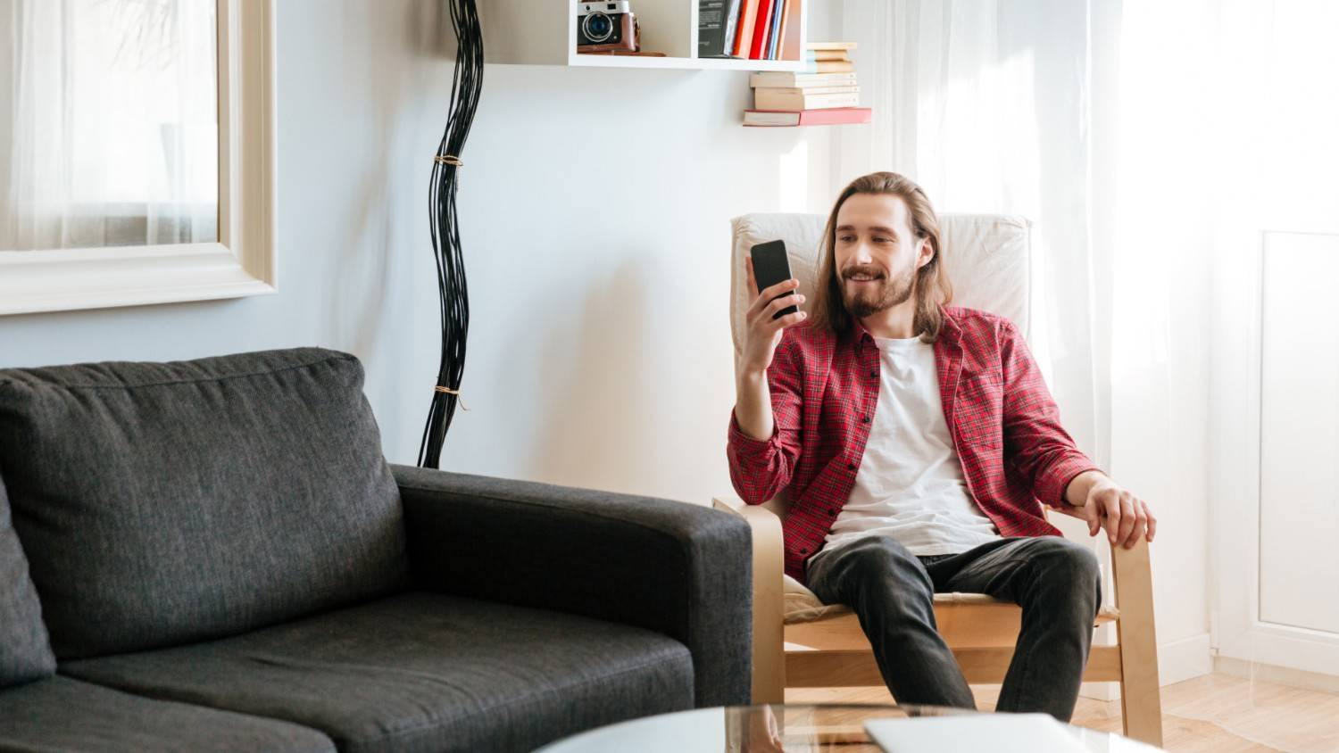A young man with long brown hair wears a white T-shirt underneath an open red shirt while sitting on a chair and reading an article on his phone about how to streamline automotive payments
